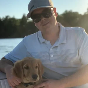 Man sitting by the water with a golden retriever puppy, wearing sunglasses and a light blue polo shirt, representing NHH Roofing Plus team member Brian Anderson, Project Estimator.